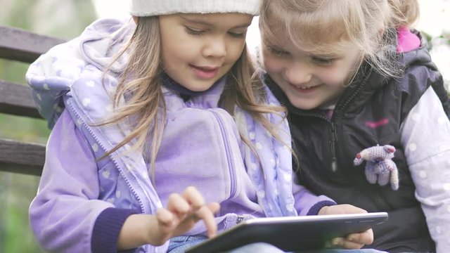 Two little girls with tablet computer sits on the bench in garden