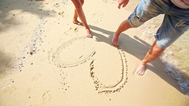 closeup caucasian girl guy feet barefoot draw heart on white wet sand on beach at wave edge