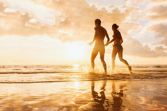 Happy cheerful couple running to the sea, water splashes, sunset