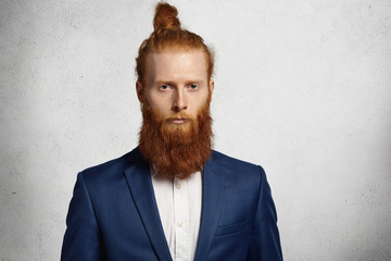 Isolated studio shot of young handsome redhead hipster man wearing trendy jacket looking serious and pensive at camera. Fashionable Caucasian businessman with stylish hair knot standing in office