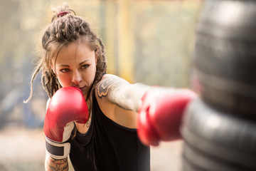 Woman boxer with tatto arms at workout. Young woman is boxing punching bag from car tires. At the head of the girl dreadlocks. Toned photo. - Powered by Adobe