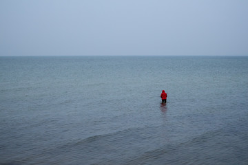Ansicht eines einsamen Anglers von hinten, der im flachen ruhigen Wasser steht um Fische zu fangen, Fischer mit roter Jacke fischt mit Angel im Meer