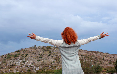 Red-haired woman standing and looking at the mountains, cloudy.