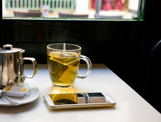 Cup of hot tea, metal tea pot and a smartphone on a table by the window in a city cafe, cozy calm atmosphere