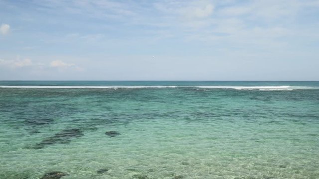 Airplane landing on island Bali airport under blue sea with waves on the horizon. Cinematic shot of overhead flying aircraft with beautiful ocean on cloudy sky background. Focal length 28mm