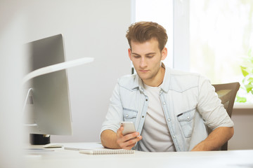 Portrait of handsome young businessman sitting at office table with personal computer and writing on mobile phone. Communication concept