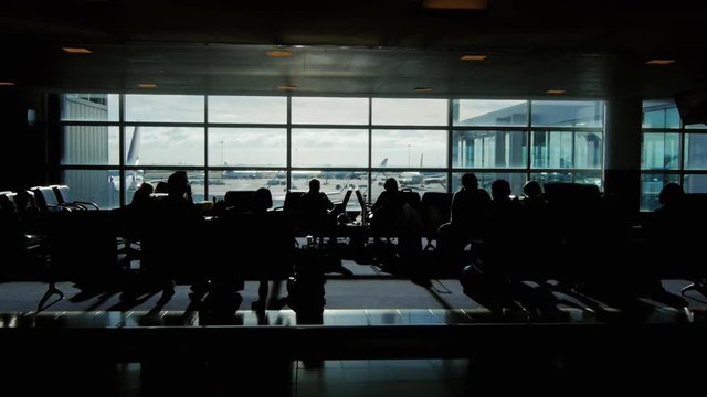 Tracking shot of a large airport terminal. silhouettes of passengers waiting for their flight. Behind the window stand airliners