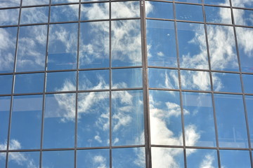 Glass wall with multiple panes held together by metal frame reflecting blue sky with white clouds.