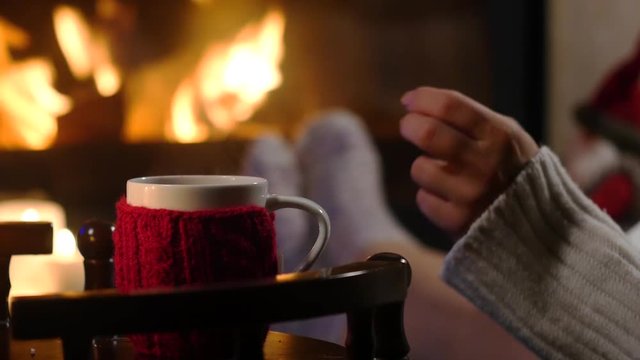 Woman is sitting with cup of hot drink and book near the fireplace