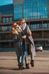 Friends walking together. Two beautiful women walking along the street holding coffee smiling and hugging.