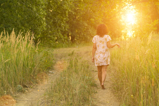 Adult brunette woman in a dress walking on a meadow path in the sunlight, view from the back
