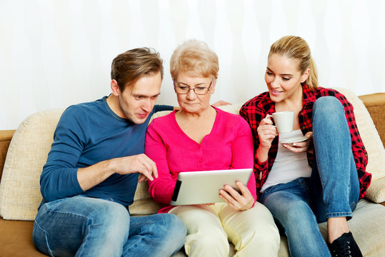 Young Couple With Old Woman Sitting On Couch And Watching Something On Tablet