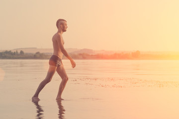 Fototapeta premium Adult man with a mohawk on his head and black shorts walking on water at sunset or sunrise