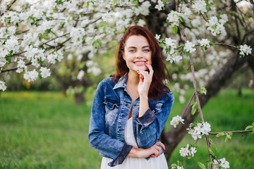 Young pretty smiling caucasian woman in jeans jacket standing near blooming spring tree. Youth, freshness, beauty, happiness concept.