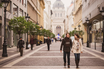 Christmas in old town. Young cheerful caucasian couple in warm cozy clothes walking in city centre. - obrazy, fototapety, plakaty