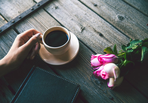 Female hand with a cup of coffee, book and flowers on wooden background. Flowers, break, work