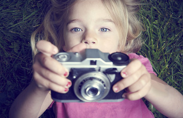 Portrait of little girl taking picture using vintage old retro film camera, lying on grass background © Petr Bonek