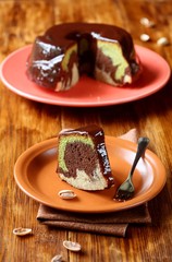 Piece of Marble Pistachio, Chocolate and Vanilla Cake covered with chocolate glaze, on brown plate and brown wooden table.
