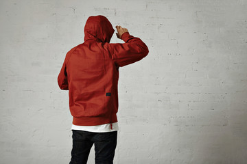 Back shot of a man in black jeans, white t-shirt and red parka putting on his hood against white wall background © BublikHaus