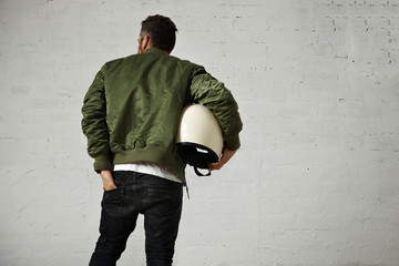 A man in khaki bomber jacket with a motorcycle helmet under his arm reaches into the back pocket of...