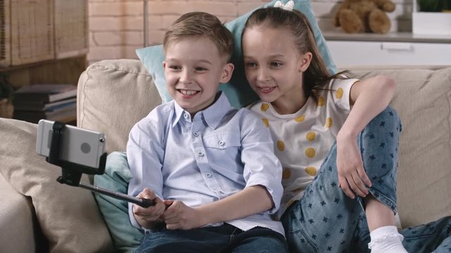 Cute little boy and girl sitting on sofa and smiling at smart phone camera while doing selfie with stick