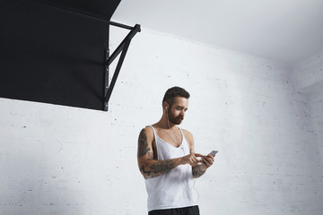 Strong tattooed athlete with smartphone and earphones chooses music playlist before workout, standing next to pull bar in fitness center