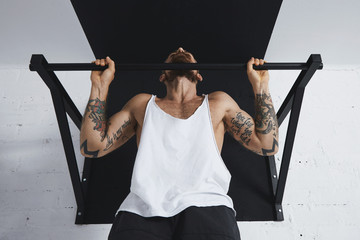 Front view on muscular male athlete in white blank tank t-shirt showing calisthenic moves Pull up with normal classic wide grip on pullbar, holding on top