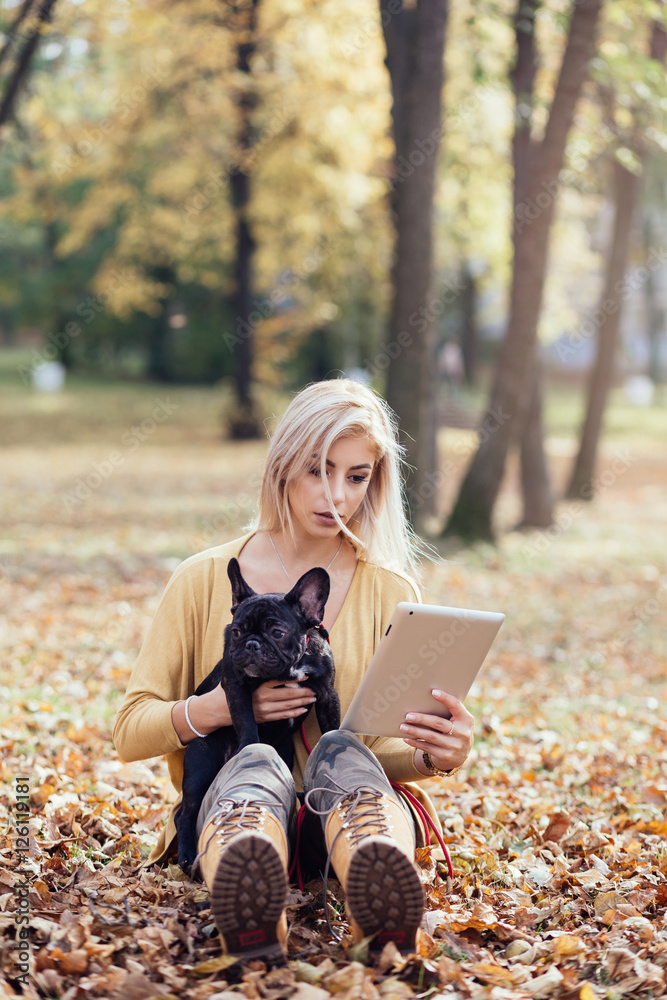 Poster beautiful and happy woman with tablet enjoying in autumn park with her adorable french bulldog.