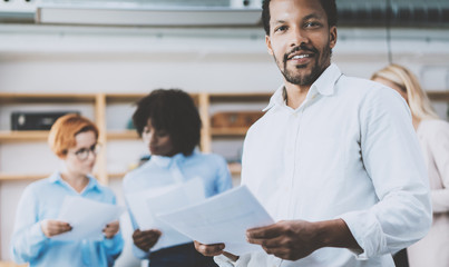 Fototapeta premium Dark skinned entrepreneur holding papers in hands and smiling at the camera.Teamwork concept in modern office. Horizontal,blurred background.