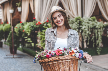 Enjoying her look. Beautiful young woman leaning at her bicycle with basket full of flowers and smiling while standing outdoors.