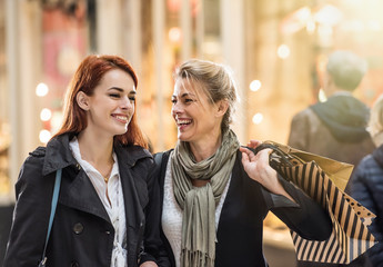 In the city, focus on a laughing mother and her daughter holding shopping bags and walking in the street. Lights of the shops at the background. Shot with flare