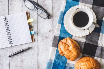 Top view of Coffee cup with bread, tablecloth, notebook, pen and glasses on wooden table background