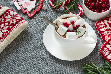 Hot chocolate with marshmallow, knitted gloves and hat, cranberries and rosemary on grey background
