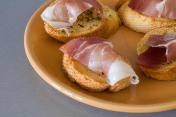Four bruschettas with prosciutto and basil on orange plate.
