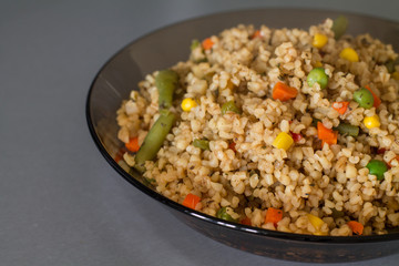 Bulgur with different vegetables, dried herb and soy sauce in a bowl on gray kitchen table.