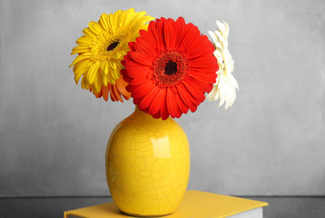 Book and yellow vase with bouquet of beautiful gerbera flowers on grey background