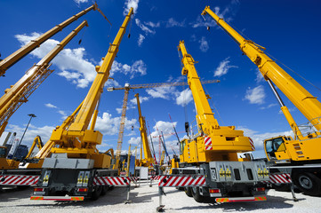 Mobile construction cranes with yellow telescopic arms and big tower cranes in sunny day with white clouds and deep blue sky on background, heavy industry  - 126170149