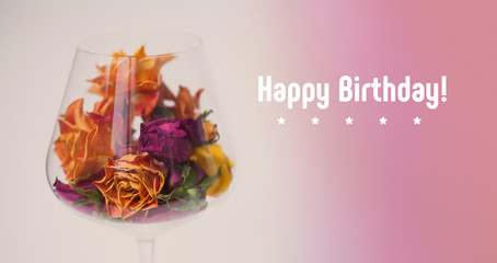 Happy birthday card decorated dried rose flowers in wine glass, pink violet gradient background. orange yellow pink flower petals macro view, copy space, soft focus