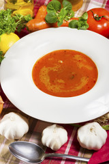 Red fish soup in a white plate beside fresh vegetables.
