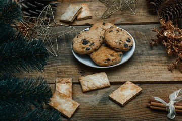 Fototapeta premium Chocolate cookies in a white plate on wooden background. Near cones and cinnamon stick, roughly. View from above. The theme of the new year holiday