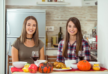 Two smiling young women are sitting at dining table and having Thanksgiving dinner with their family.