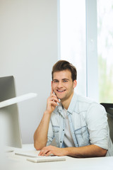 A young programmer is looking at the camera. The graphic designer is thinking about the future projects. The young guy is sitting at the computer in the office with the windows behind his back