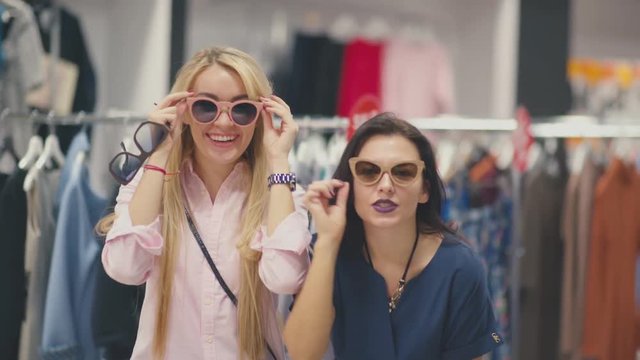 Portrait of two funny friends. Two young women trying on sunglasses in store and play the fool in front of the mirror. Brunette and blonde - two friends try on glasses and having fun.