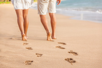Beach couple relaxing at sunset walking barefoot. Focus on footprints in golden sand. Closeup of legs. Romantic beach vacation holidays. Young people from behind walking away towards happiness.