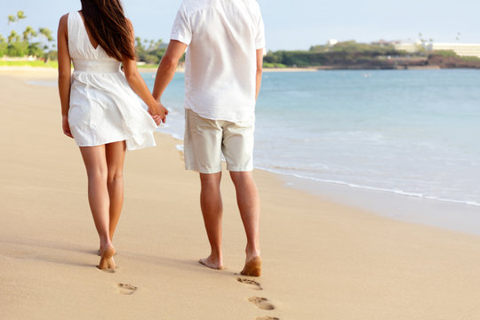 Honeymoon Barefoot Couple Holding Hands Walking On Romantic Beach Vacation Holidays Leaving Footprints In The Golden Sand With Feet. Young People From Behind In White Shorts And Dress Beachwear.