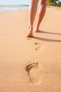 Beach sand footprints woman feet walking barefoot. Travel girl relaxing walking on golden sand beach leaving footprints in the sand. Closeup detail of female feet and legs at sunset.