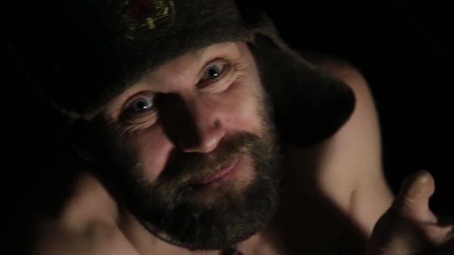 scary evil sinister bearded man with smirk, jumps abruptly from the darkness. strange Russian man with a naked torso and a woolen hat