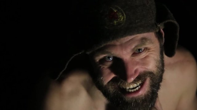scary evil sinister bearded man with smirk, angrily looks into the camera. strange Russian man with a naked torso and a woolen hat