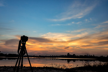 Naklejka premium silhouette of camera on tripod with sunset sky on lake.