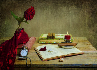 Still life with red rose wrapped in silky scarf placed with old book, vintage box of cigar, candle,shell and pocket watch on rustic wooden table. 
